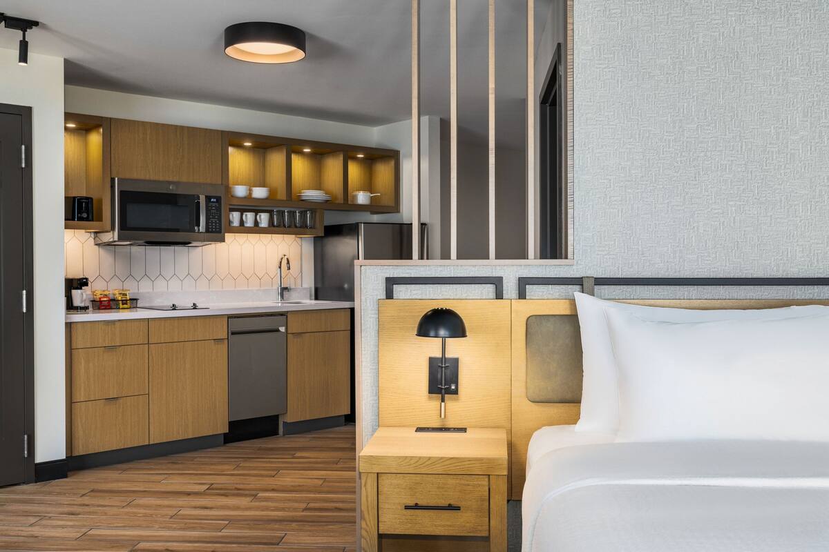 Una habitación de hotel moderna con piso de madera, una cama con sábanas blancas, una mesita de noche con lámpara, microondas integrado, y una pequeña cocina con gabinetes de madera y lavavajillas.