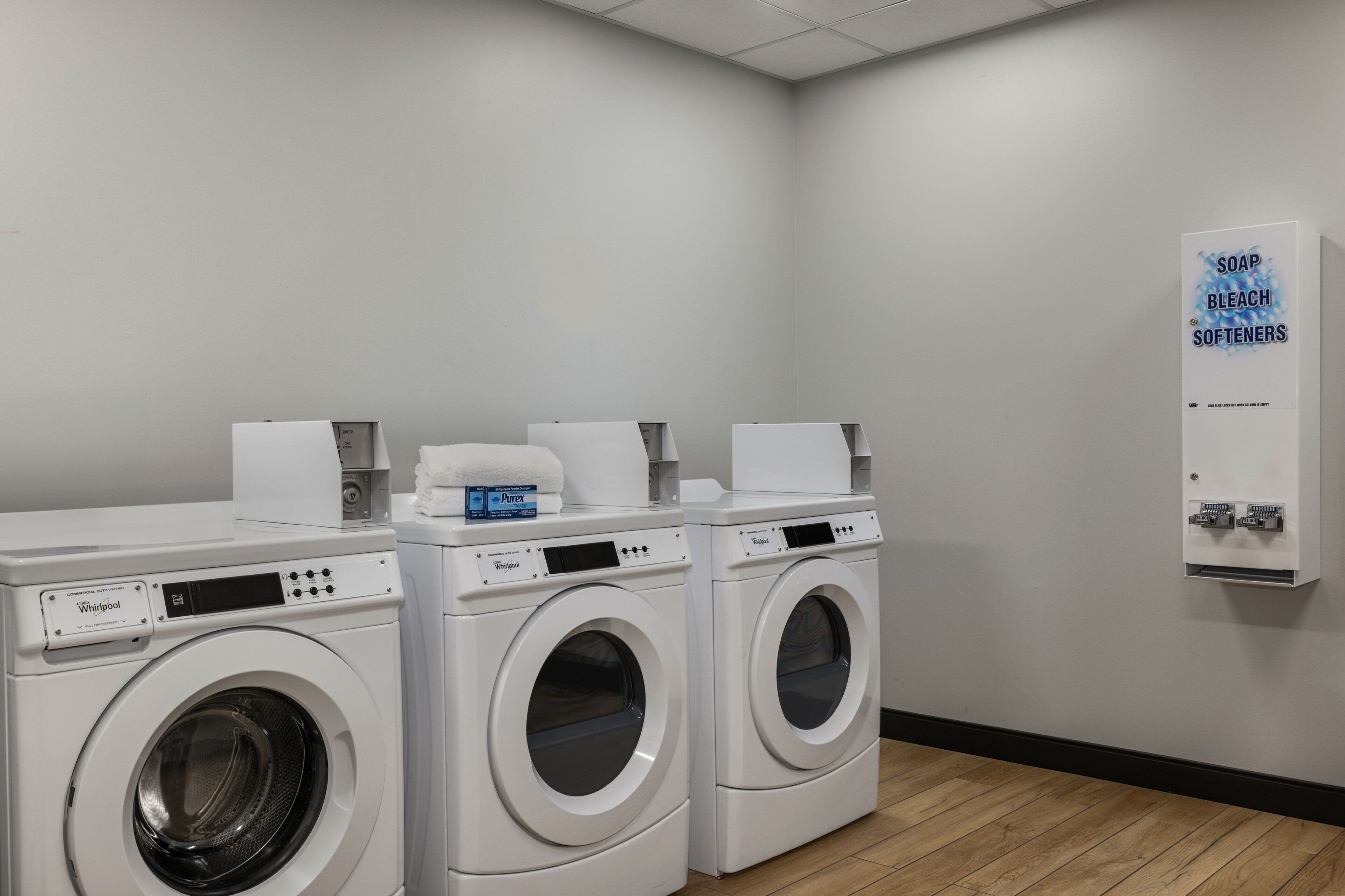 Une buanderie équipée de deux lave-linge Whirlpool à chargement frontal, d’un distributeur de savon et d’eau de Javel fixé au mur, et d’une boîte de lessive Purex.