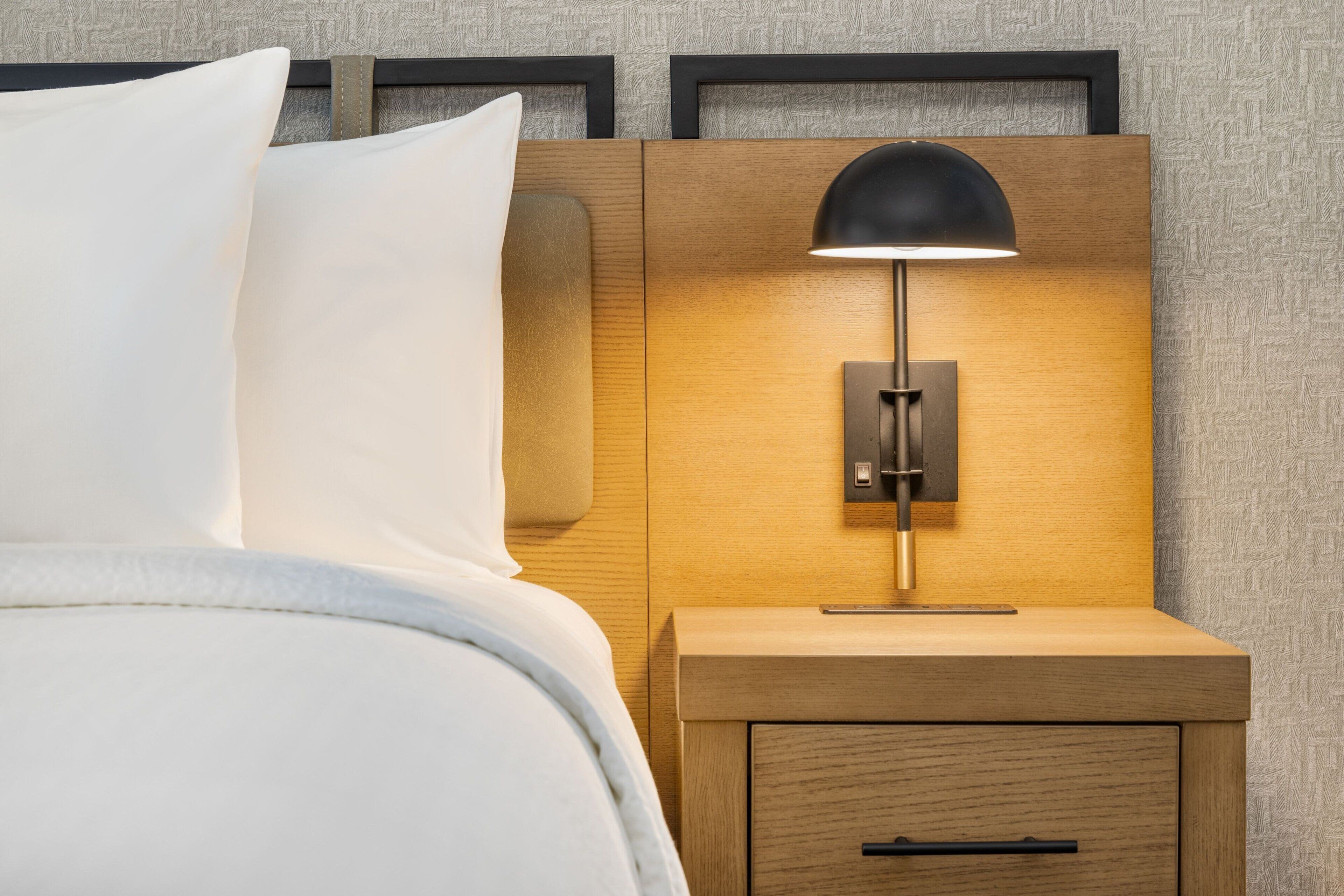 Un lit bien fait, avec du linge de lit blanc et une table de chevet en bois surmontée d’une lampe noire.