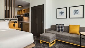A hotel room with a bed, a gray sofa, an ottoman, a small table, a kitchenette, and two framed pictures on the wall. - Towneplace Suites By Marriott Memphis Germantown (Germantown)