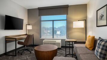 A hotel room with a flat-screen TV, a desk with a chair, a sofa with a plaid throw pillow, a round wooden coffee table, and a large window with a view of buildings and trees.