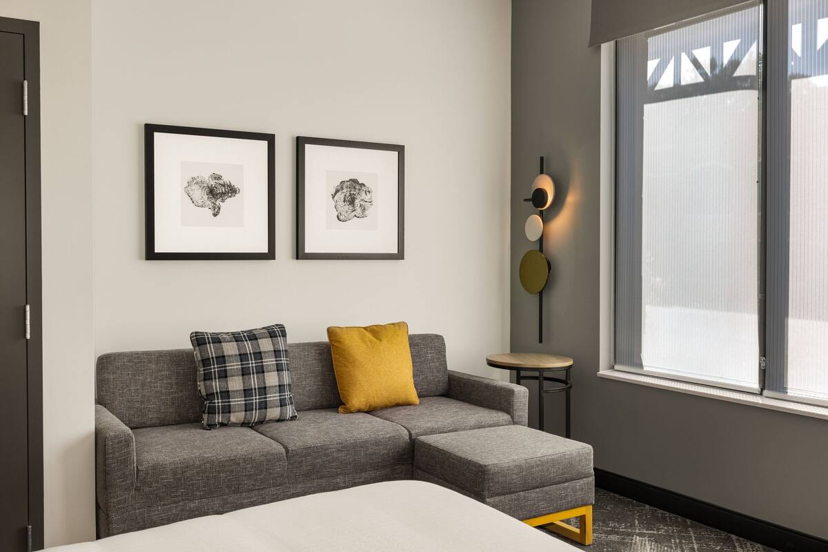 Una habitación de hotel moderna con un sofá seccional gris, dos cuadros en la pared, una mesita redonda con una lámpara y un amplio ventanal con persianas.