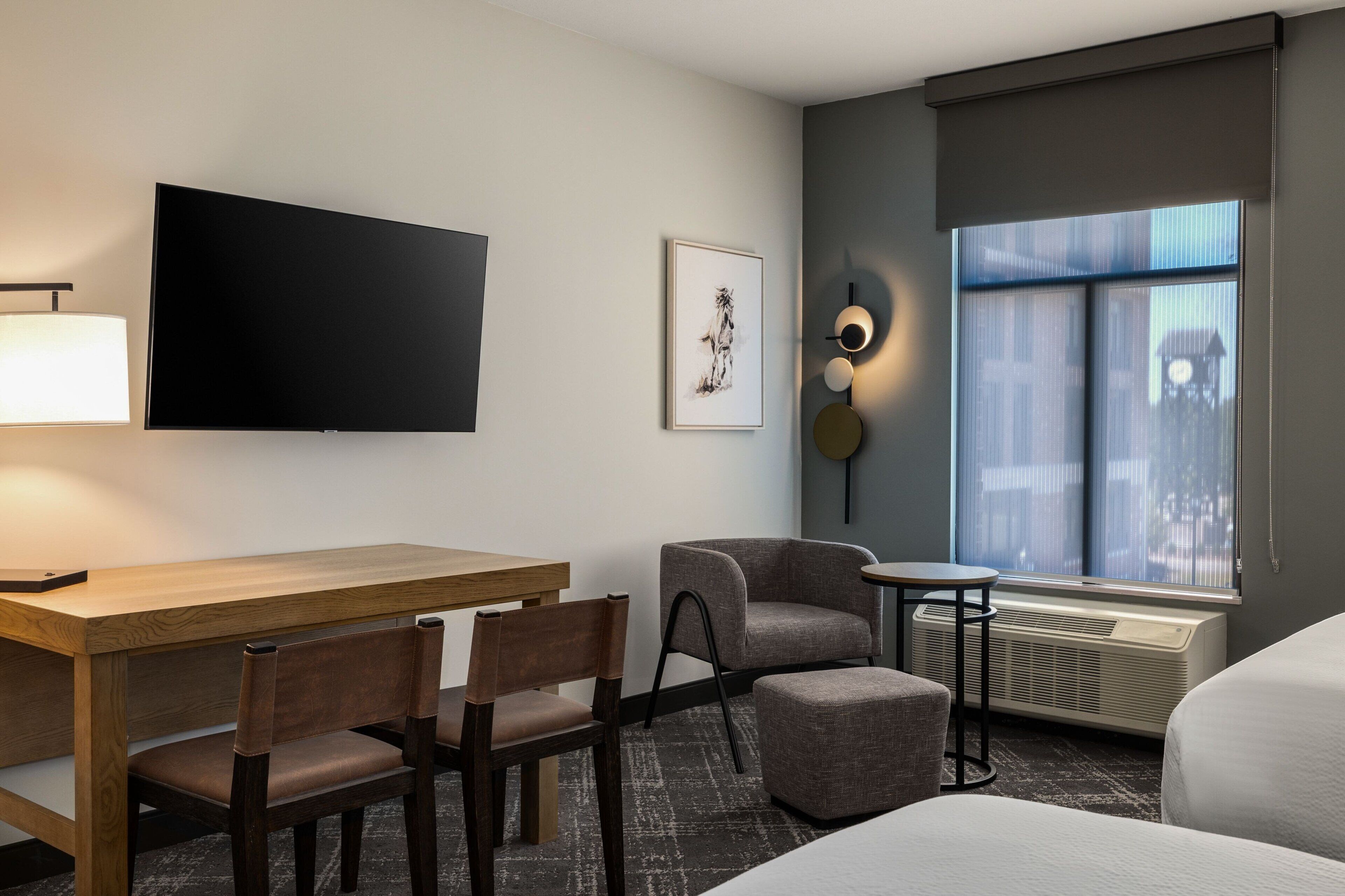 A hotel room with a wooden desk, two chairs, a small round table, a sofa, a TV mounted on the wall, a framed artwork, a wall-mounted light fixture, and a large window with a view of buildings.