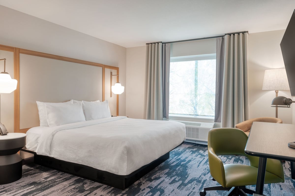 Habitación de hotel con una cama grande, un escritorio, una silla, una lámpara y un ventanal con cortinas.