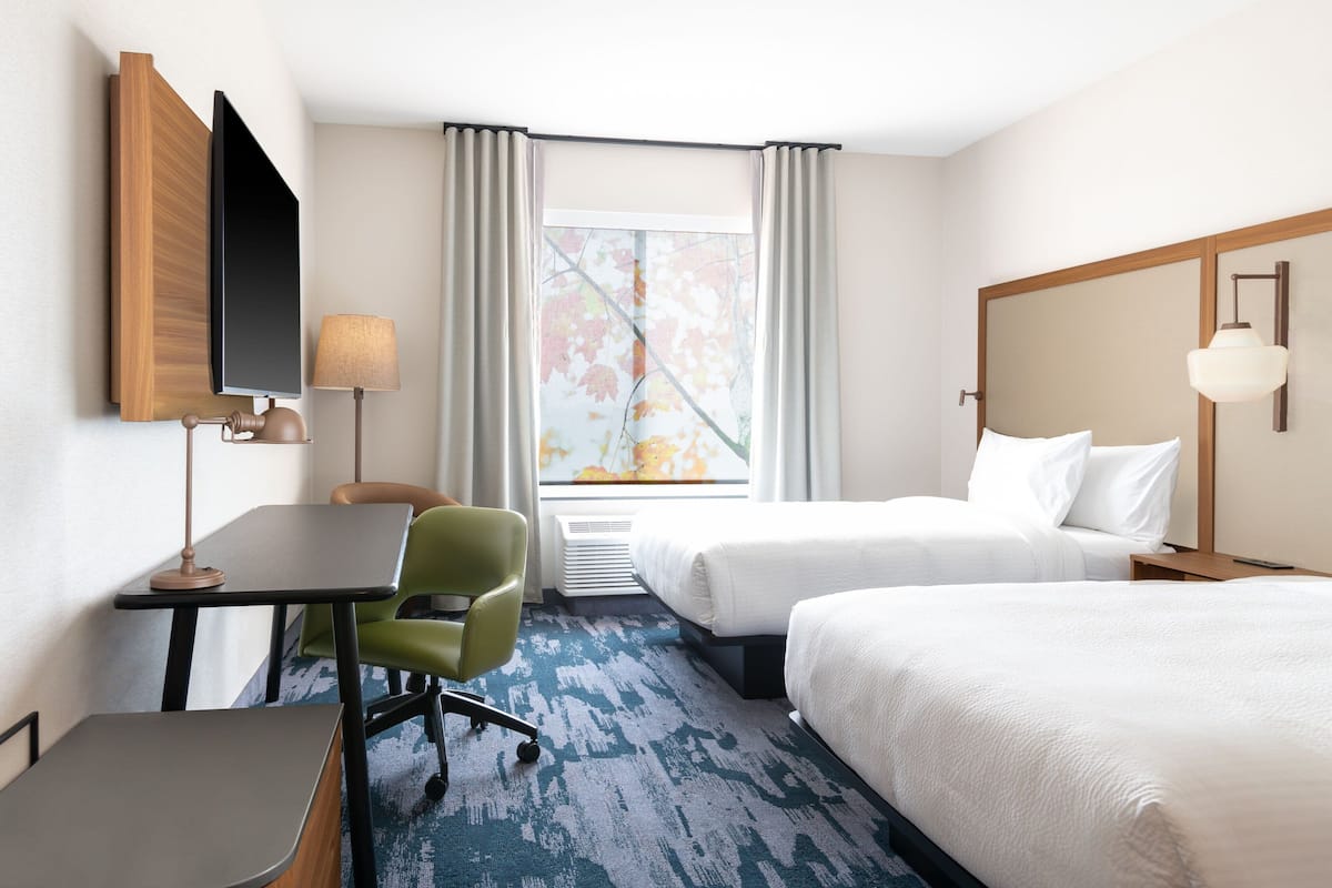 A hotel room with a desk, two beds, a chair, a lamp, and a window with curtains.