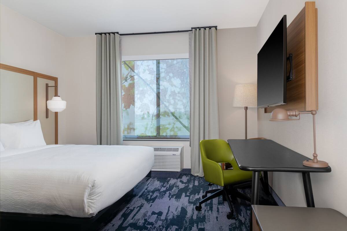 A hotel room with a bed, a desk, a chair, a TV, and a window with curtains.