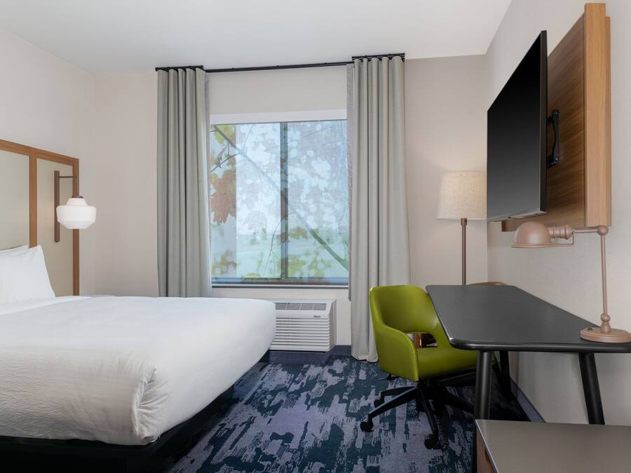 A hotel room with a bed, a desk, a chair, a TV, and a window with curtains.