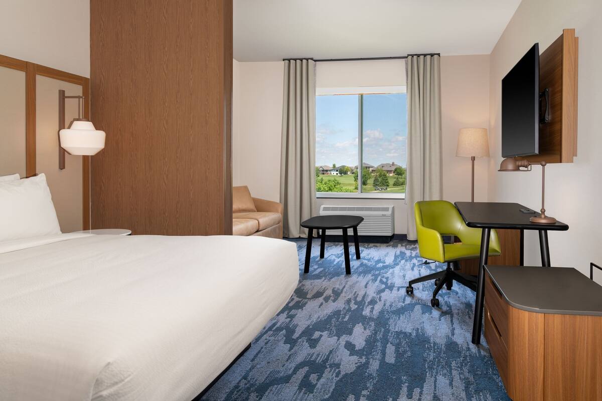 A hotel room with a large bed, a desk, a chair, a TV, and a window with a view of houses.