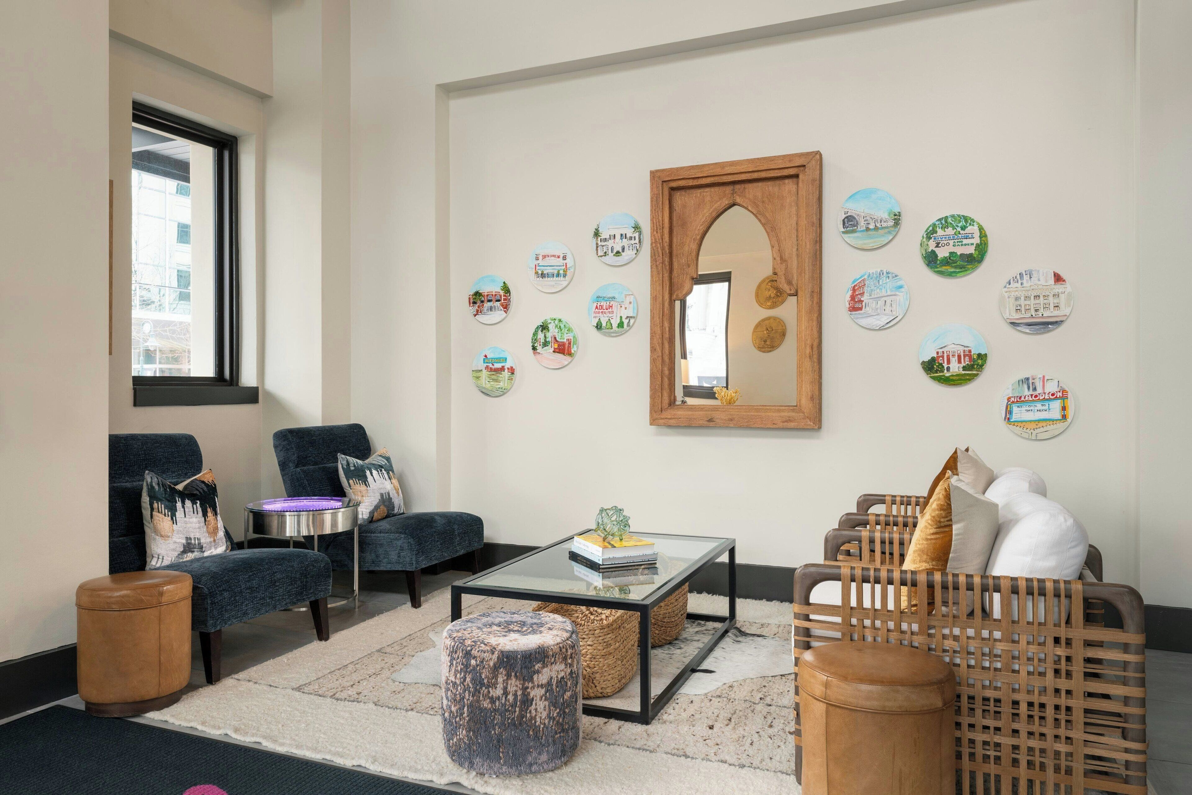 A modern living room with a glass coffee table, two blue armchairs, a wooden sofa, and a decorative mirror.