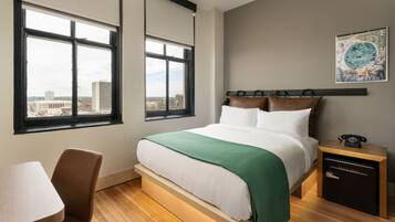 A hotel room with a bed, a desk, and a view of the cityscape through large windows.