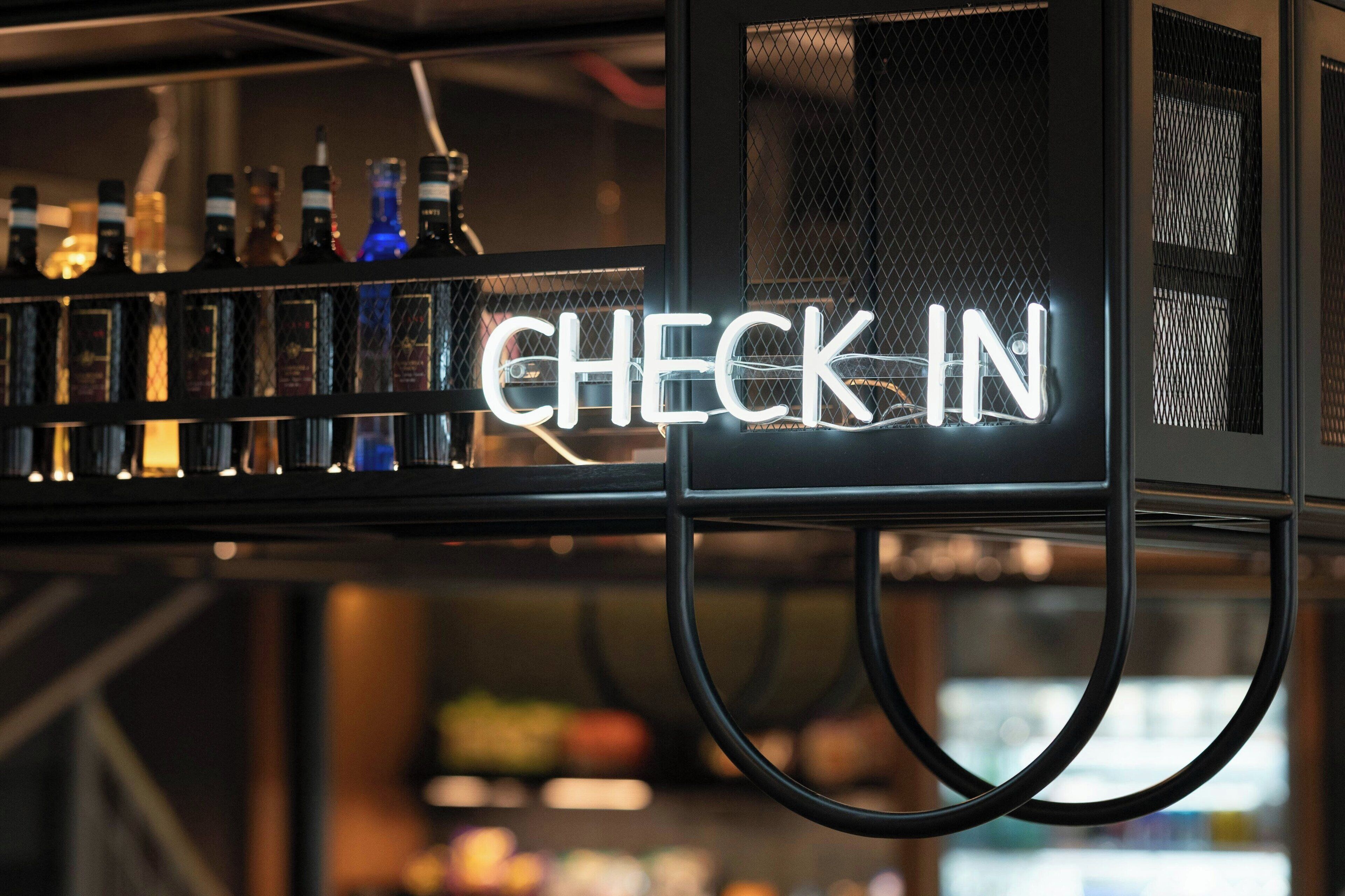 Uma área de bar com um letreiro de neon 'CHECK IN' e prateleiras cheias de garrafas de bebidas.