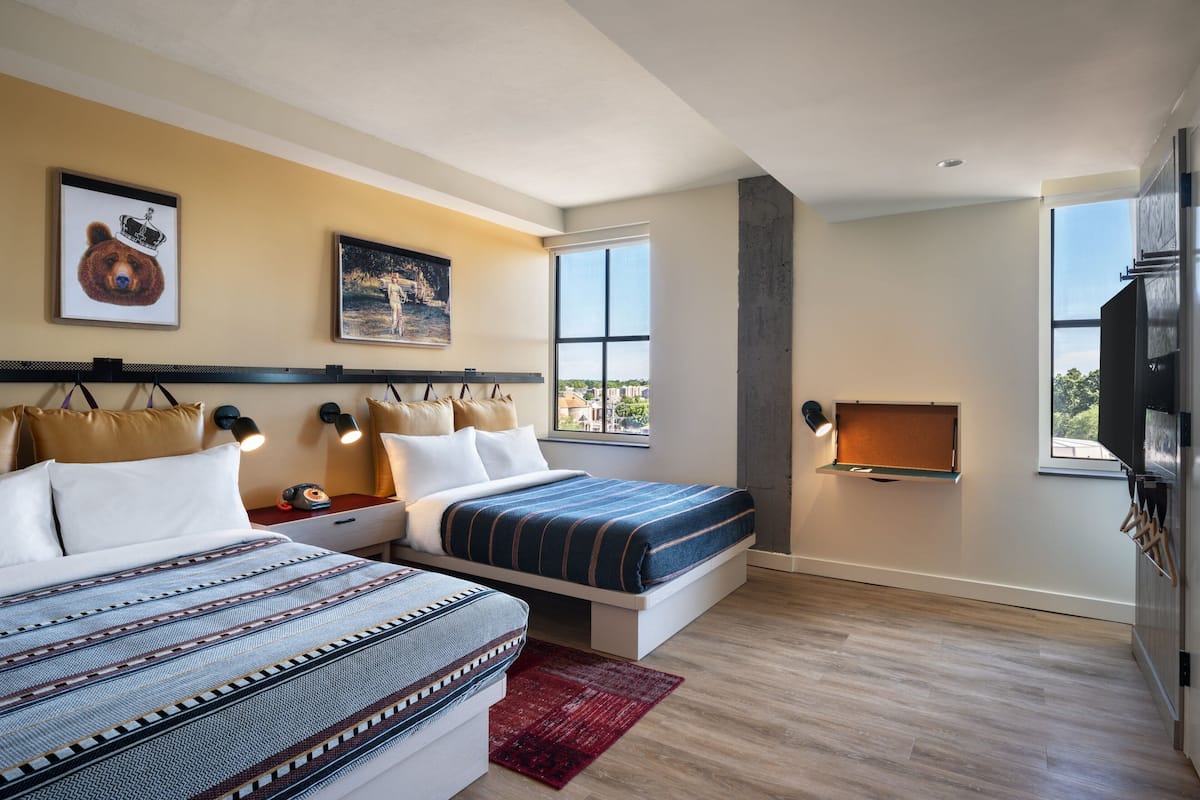 A modern hotel room with two beds, a large window with a city view, a mounted TV, and artwork on the walls.