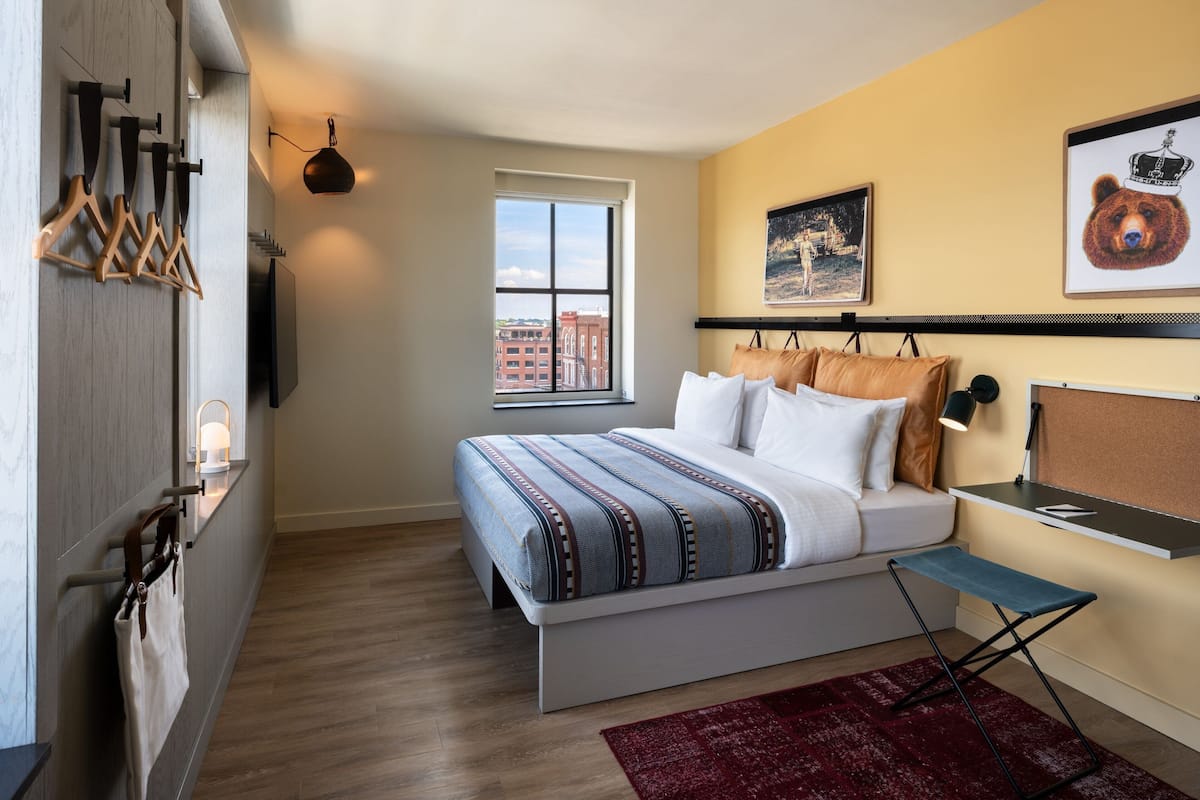 A bedroom with a bed, a window, a wall-mounted TV, and a red rug.