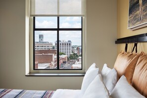 Quarto de hotel com uma janela ampla que oferece vista para a cidade, uma cama com roupa branca e cabeceira com vários travesseiros.