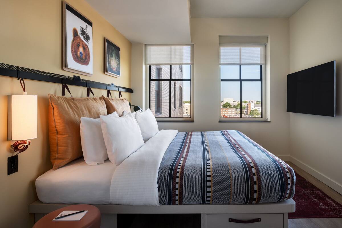 A hotel room with a large bed, two windows, a mounted TV, and artwork on the wall.