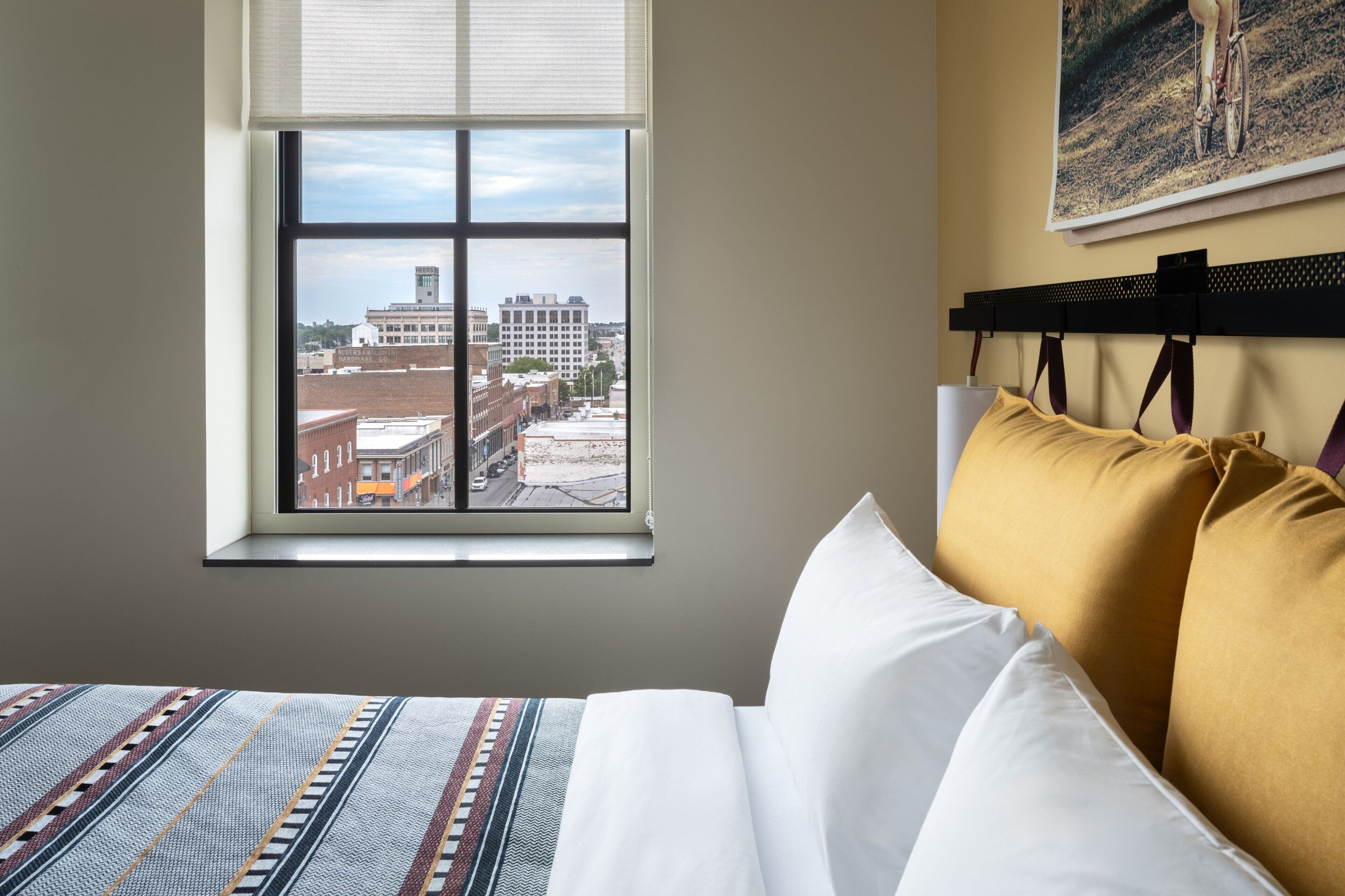 A hotel room with a large window offering a city view, a neatly made bed, and a headboard with multiple pillows.