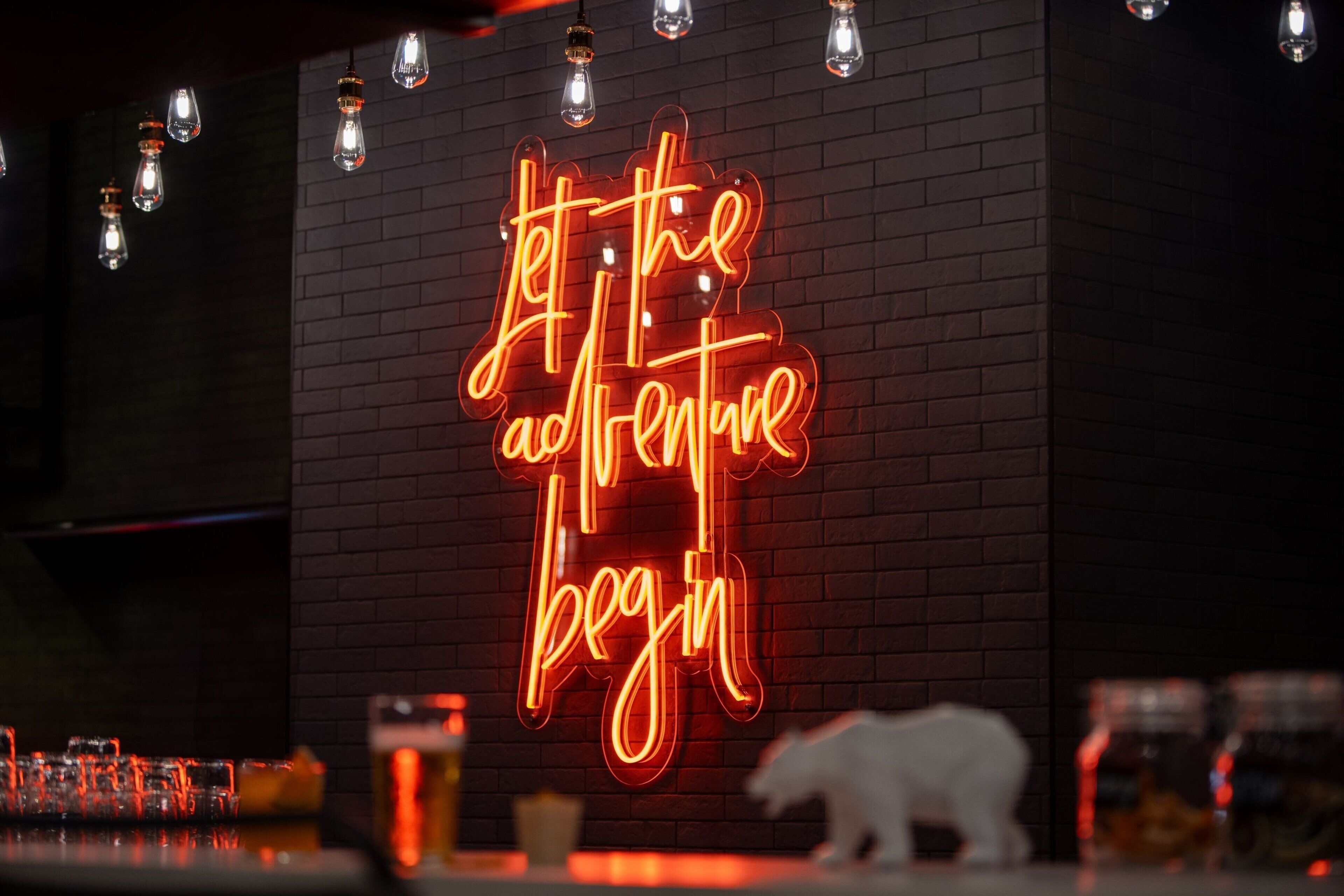 A neon sign with the text "Let the adventure begin" on a brick wall.