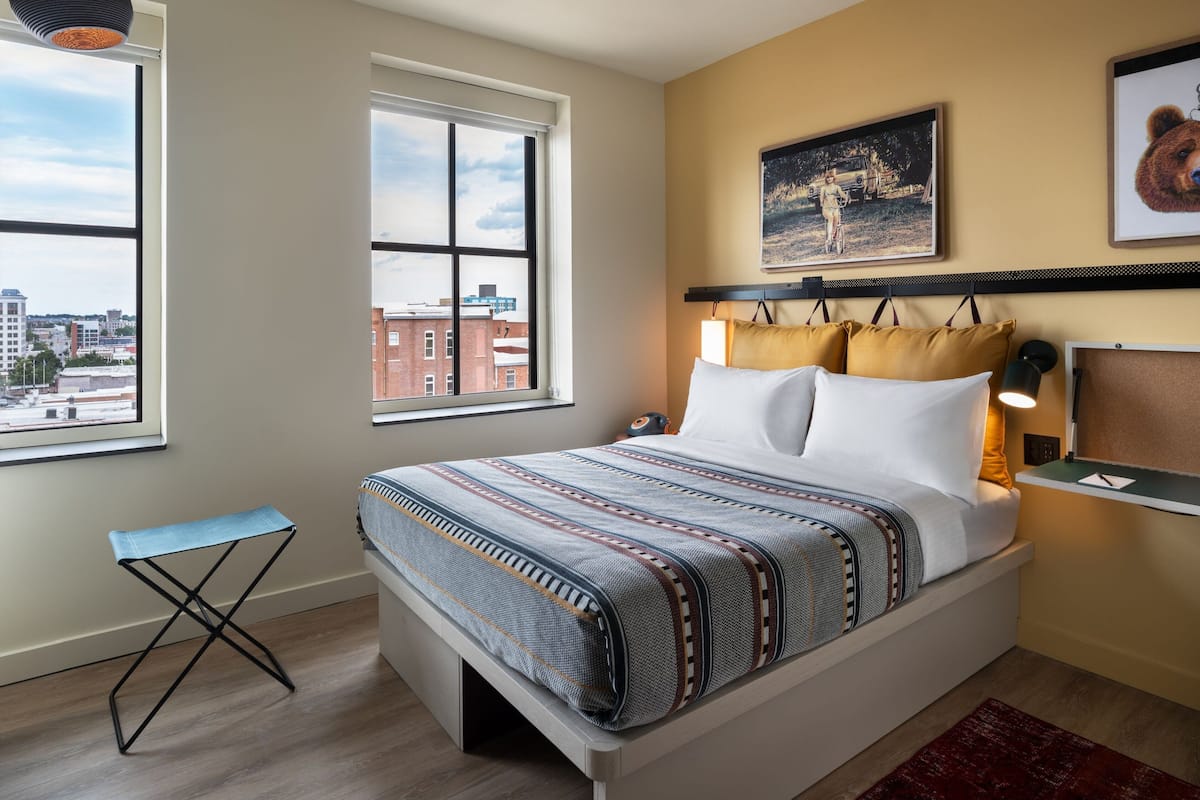 A bedroom with a bed, a window, a small table, and a lamp.