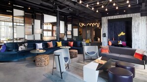 A modern lounge area with a blue sofa, white armchairs, and a dark sofa with colorful cushions. There are round tables and a neon sign on the wall. - Aloft by Marriott McAllen (McAllen)