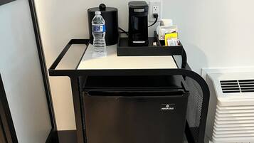 A compact minibar with a water bottle, coffee maker, and a small box, likely containing toiletries, against a white wall with a flat-screen TV mounted above.