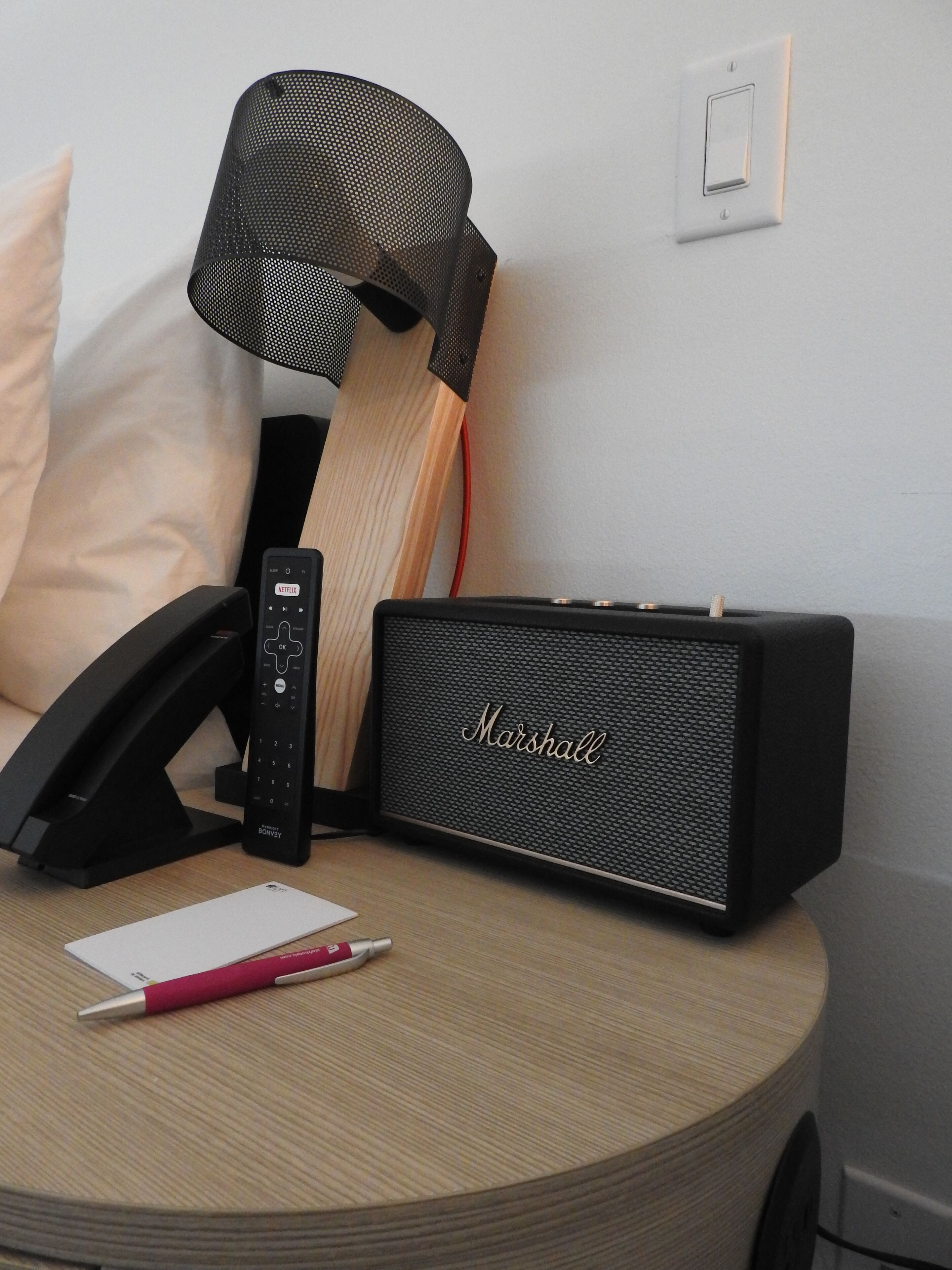 A round wooden table with a black speaker, a remote control, a book, and a pink pen.
