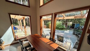 Un comedor con una amplia mesa de madera y sillas, ventanas grandes con vistas a un área boscosa y un vehículo en el exterior.