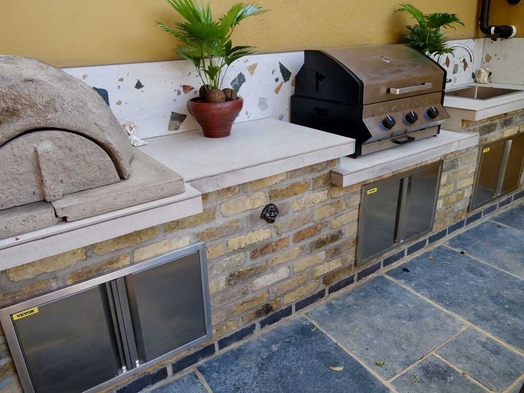 An outdoor kitchen with a stone wall, a pizza oven, a grill, and a sink.