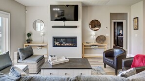 Un salon doté d’une cheminée, d’un téléviseur fixé au-dessus, et d’un coin salon confortable avec un canapé et des fauteuils.