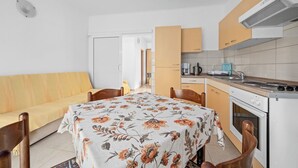 Een kleine keuken met een fornuis, spoelbak en kastjes. Er is een eettafel met stoelen en een bank in de woonkamer.