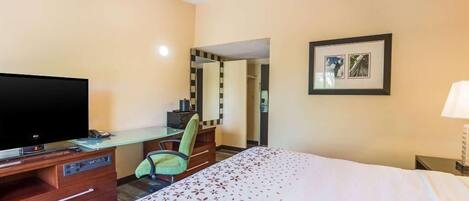 A hotel room with a bed, a desk with a computer, a television, a chair, a mirror, and a framed picture on the wall.