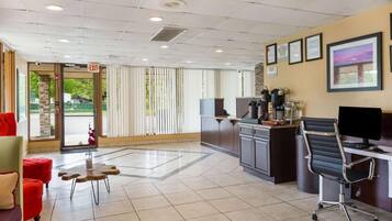Vestíbulo de hotel con mostrador de recepción, computadora, estación de café y zona de estar con un sofá rojo y una mesa de madera.