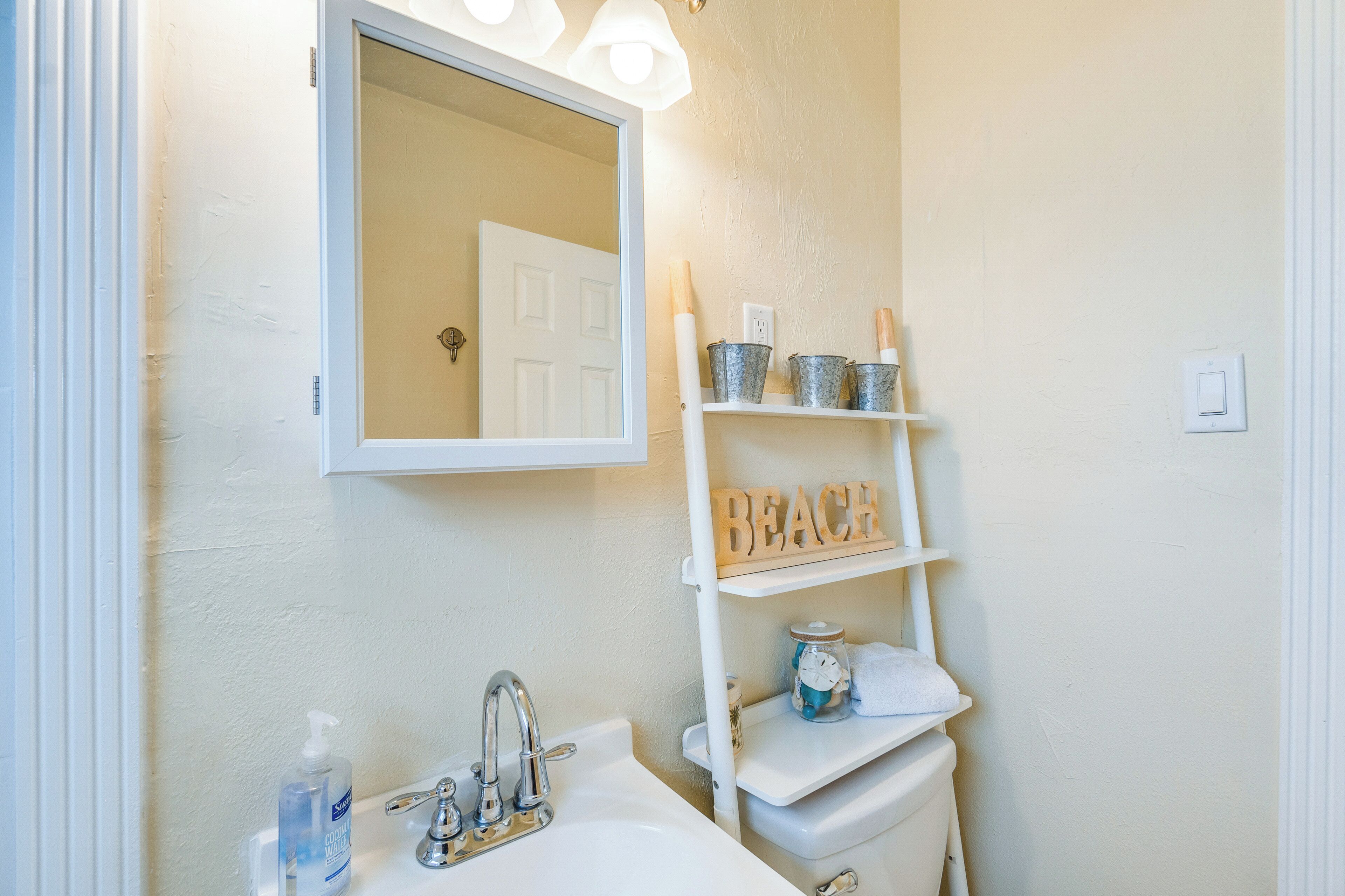 A bathroom with a white sink, a mirror above it, and a shelf with a 'BEACH' sign.