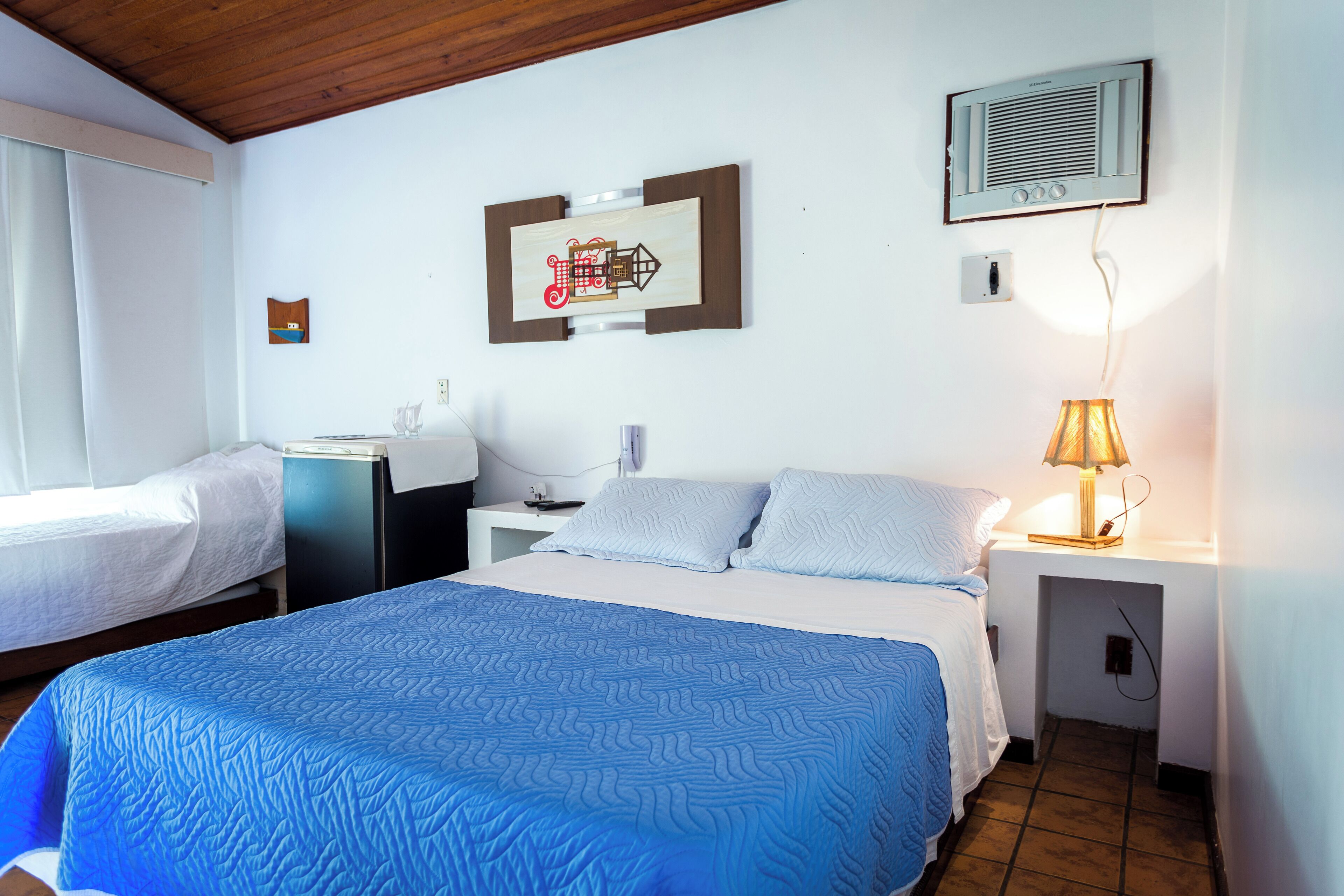 A hotel room with two single beds, a desk with a lamp, a wall-mounted air conditioner, and a framed artwork.