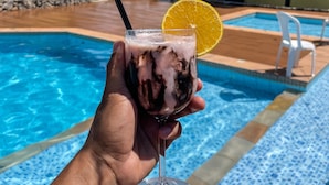 Mão segurando um copo com bebida e uma fatia de laranja, perto de uma piscina com bordas azulejadas e um edifício ao fundo.