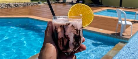 Une main tenant un verre contenant une boisson et une tranche d’orange, près d’une piscine dont les bords sont recouverts de carreaux, avec un bâtiment en arrière-plan.