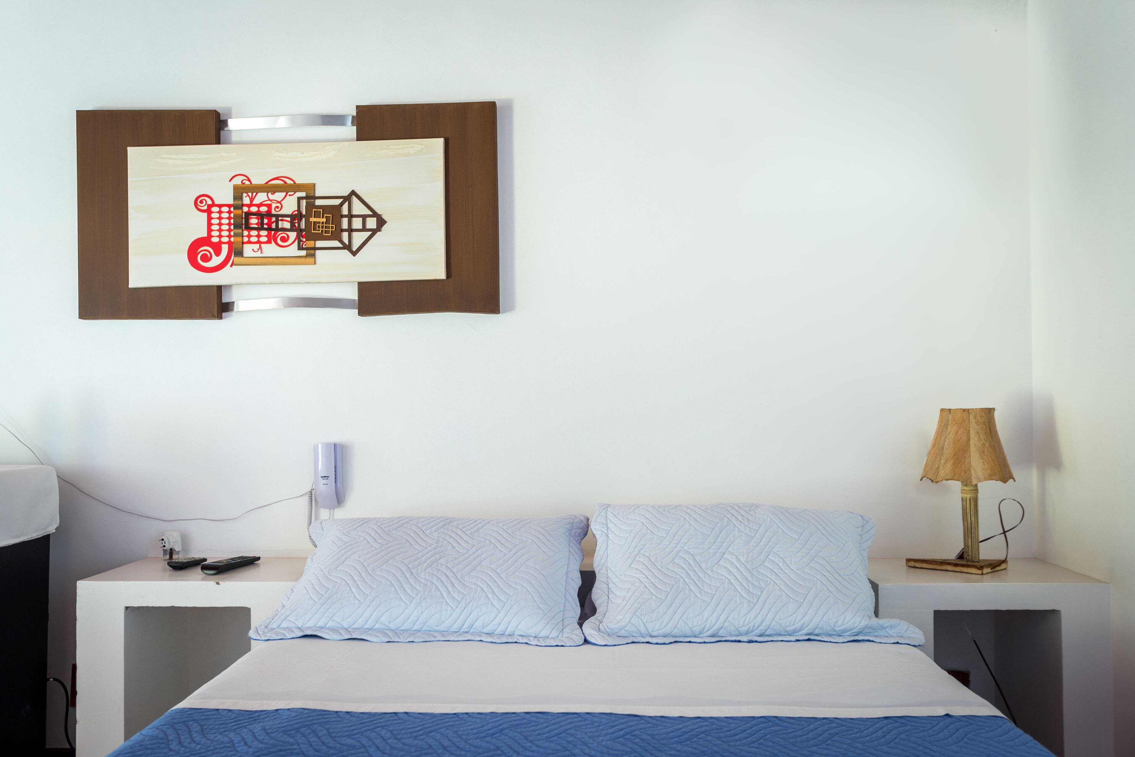 A hotel room with a bed, a bedside table with a lamp, a wall-mounted artwork, and a phone on the table.