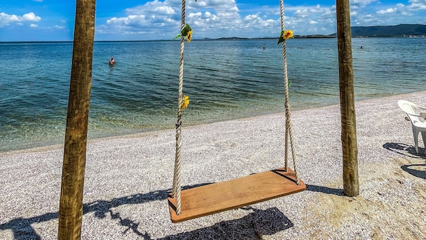 Um baloiço de madeira com uma placa decorativa, situado numa praia de seixos com vista clara para o mar e montanhas ao longe.