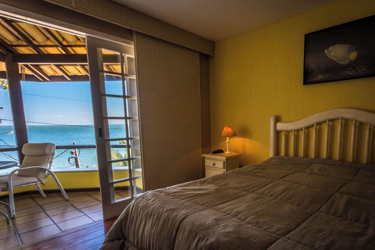 Una habitación de hotel con cama, una silla, mesita de noche, lámpara y vista al océano.