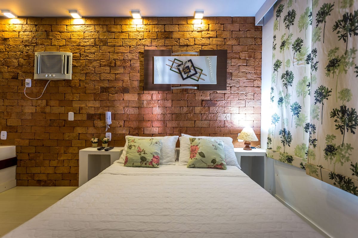 Habitación de hotel con pared de ladrillo, aire acondicionado instalado, televisor de pantalla plana, almohadas estampadas con motivos florales, mesita de noche con lámpara y cortina con diseño floral.