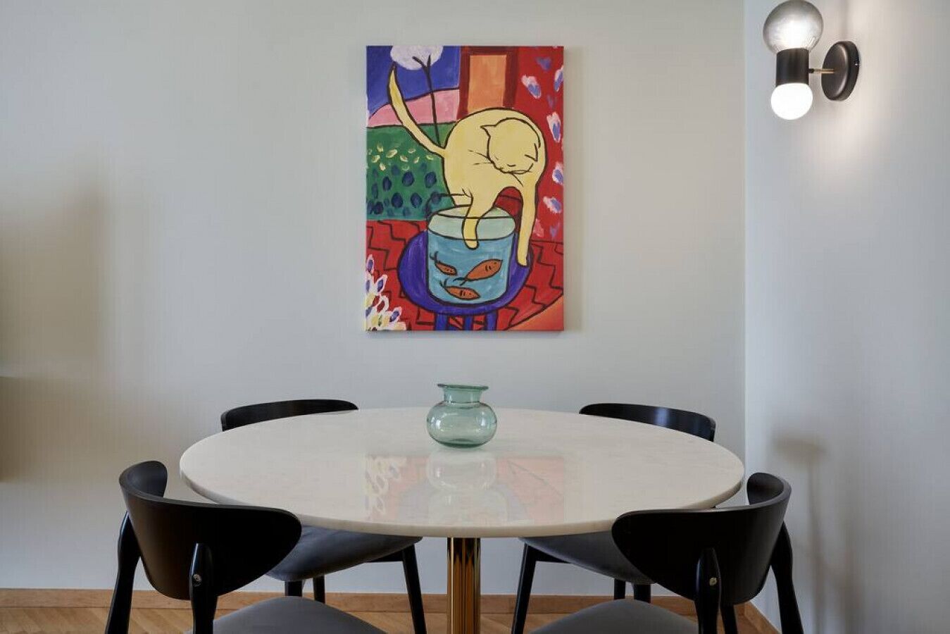 A dining area with a round table, black chairs, a glass vase, and a colorful abstract painting on the wall.