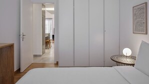 Ein Hotelzimmer mit Bett, Nachttisch mit Lampe, Schrank und Blick auf einen Essbereich.