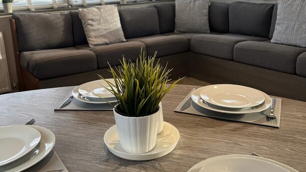 A dining area with a table set for four, a grey sofa, and a potted plant in the center. - 3 bed Stunning Caravan Situated nr Whitstable (Whitstable)