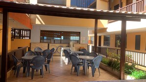 An outdoor patio with tables and chairs, a tiled floor, and a kitchen area in the background. - Clube Praiano (Cabo Frio)