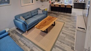 A modern living room with a blue sofa, a wooden coffee table, and a kitchen in the background. - Charming 3-bed House in Newquay (Newquay)