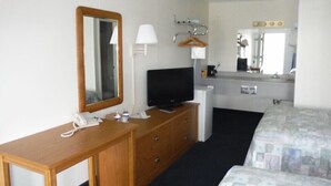 Ein Hotelzimmer mit Holzschreibtisch, Fernseher, Spiegel, Nachttisch, einem Bett mit gemusterten Bettwäsche, einer kleinen Kommode, einer Lampe und einem Kleiderhaken.