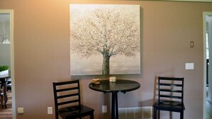 A dining area with a round table and two chairs, a painting of a tree on the wall, and a light fixture above the table.