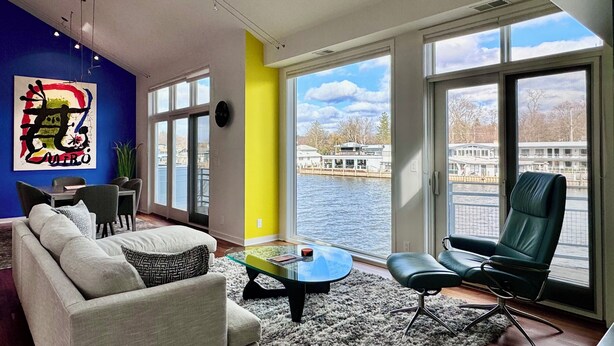 A modern living room with a sofa, armchair, and coffee table, large windows with a view of a river and boats, and a colorful abstract painting on the wall. - Stunning 3BR Condo w/6 Decks & a Boat Dock (Saugatuck)