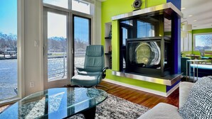 A modern living room with a glass coffee table, a comfortable chair, a fireplace, and large windows offering a view of the outdoors. - Stunning 3BR Condo w/6 Decks & a Boat Dock (Saugatuck)