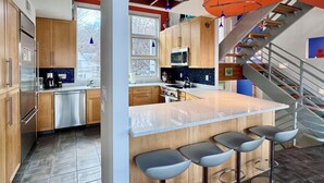 A modern kitchen with wooden cabinets, stainless steel appliances, a large island with bar stools, and a staircase with a metal railing. - Stunning 3BR Condo w/6 Decks & a Boat Dock (Saugatuck)