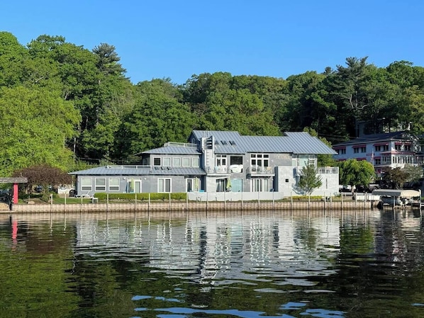 A waterfront property with a large house featuring multiple levels, a dock, and a reflection in the water.